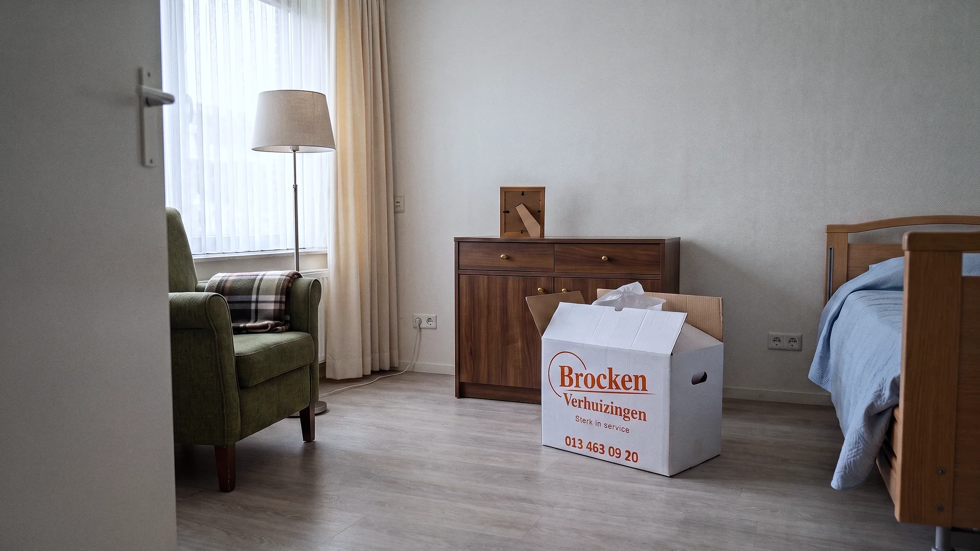 Een kamer in een verpleeghuis met een eigen houten kastje, een leunstoel en een geopende verhuisdoos.