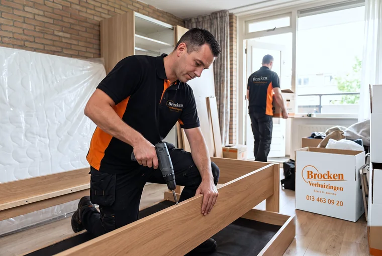 Verhuizer van Brocken Verhuizingen schroeft een houten bed uit elkaar, terwijl een collega een verhuisdoos naar buiten draagt