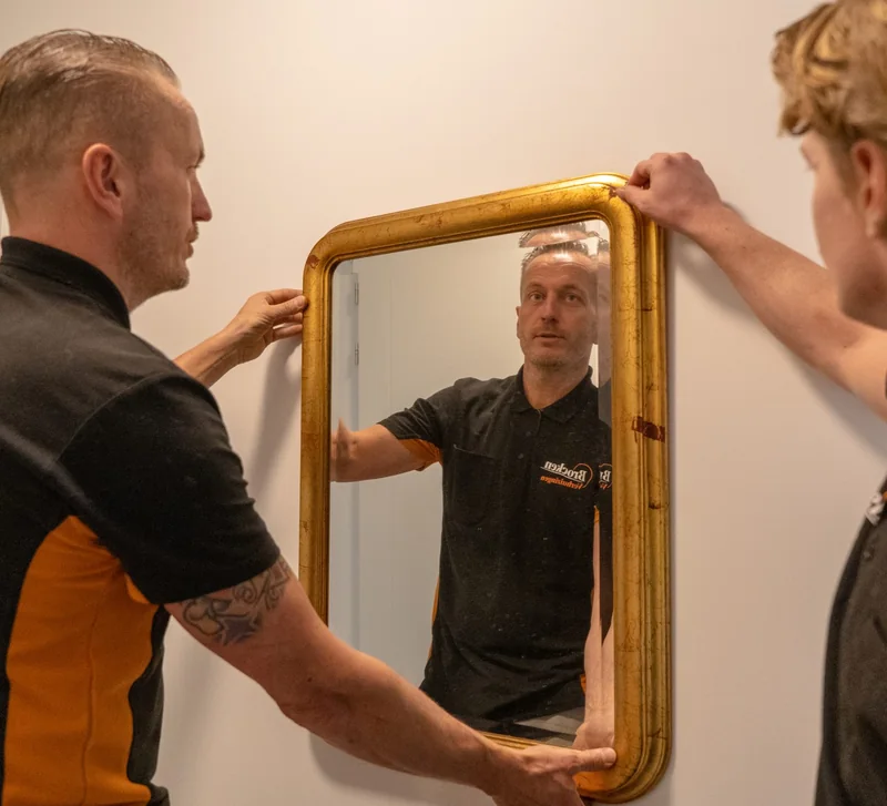 Twee verhuizers van Brocken Verhuizingen hangen een spiegel met gouden lijst op