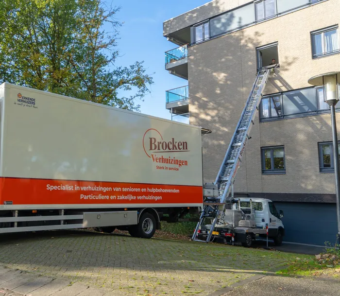 De verhuislift van Brocken Verhuizingen staat uitgeschoven tegen een appartementengebouw, naast de verhuiswagen