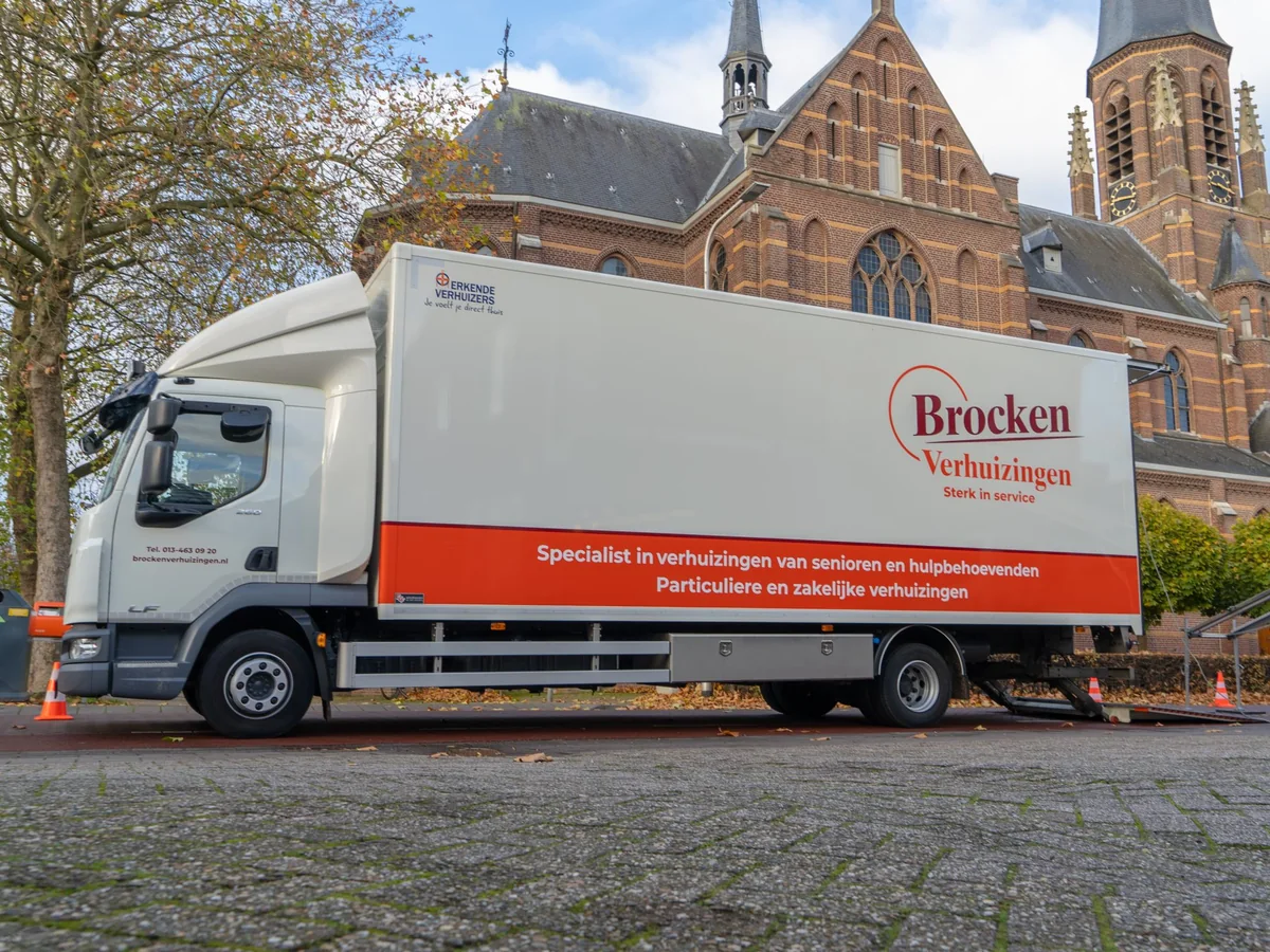 Vrachtwagen van Brocken Verhuizingen met de oranje band langs de laadbak, geparkeerd voor een kerk