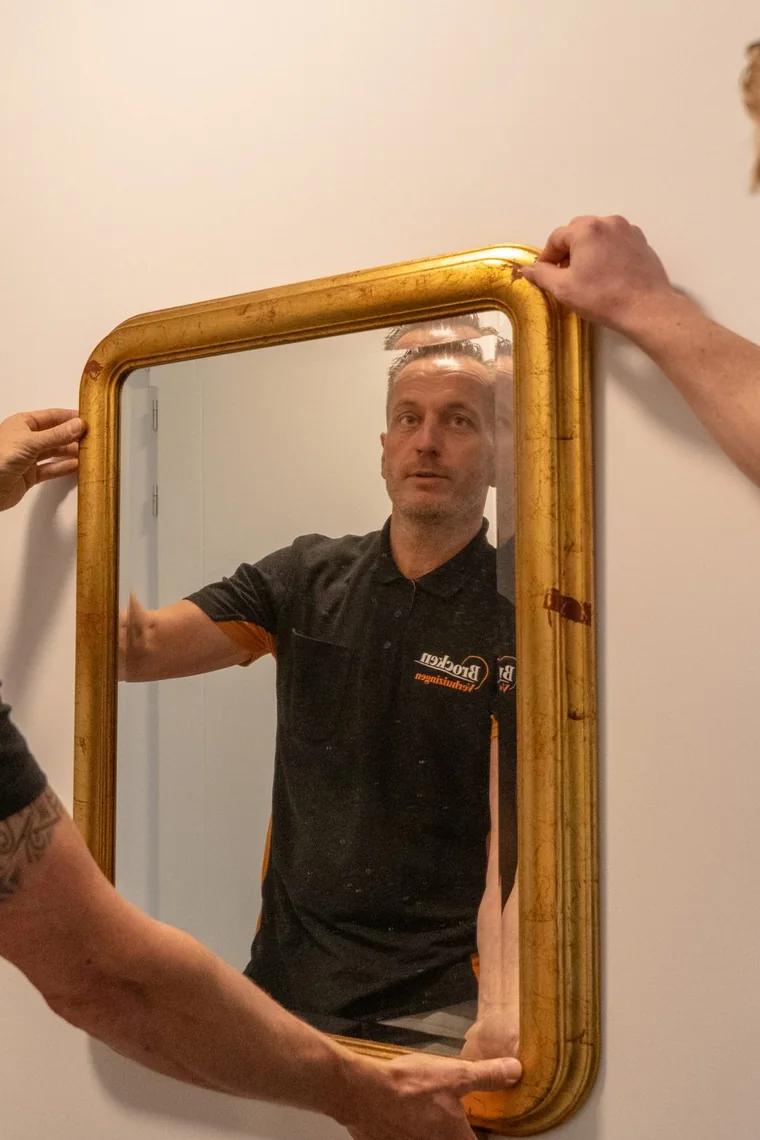 Twee verhuizers van Brocken Verhuizingen hangen een spiegel met gouden lijst aan de muur