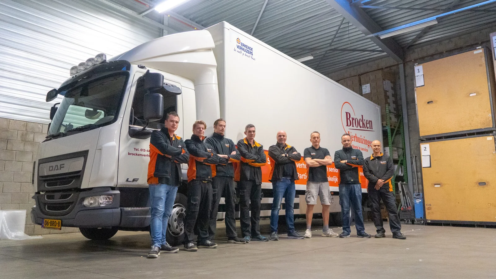 Het verhuisteam van Brocken Verhuizingen, acht verhuizers met de armen over elkaar voor de vrachtwagen in de loods
