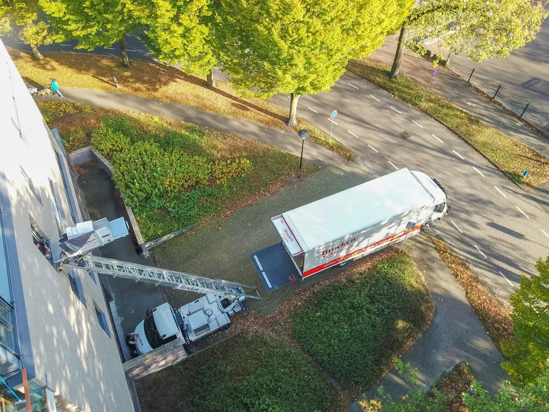 Van bovenaf gezien: de ladderlift van Brocken Verhuizingen tegen de gevel van een flat, met de verhuiswagen op het pad ernaast