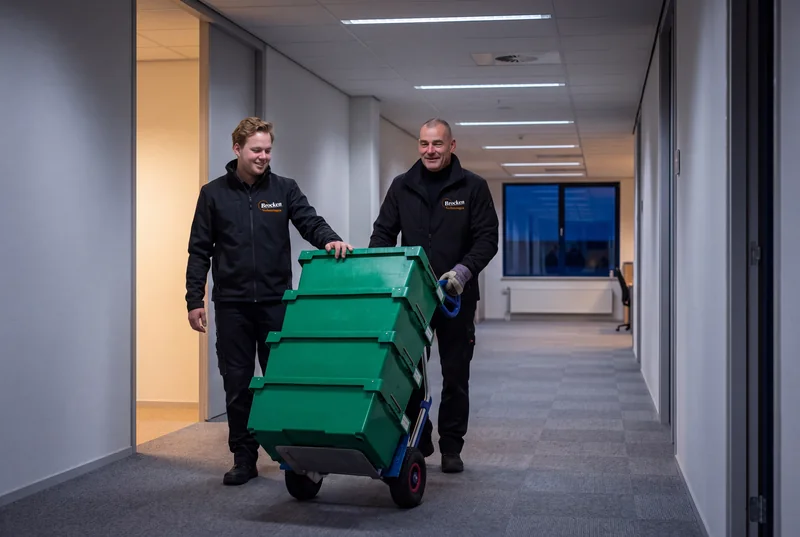 Twee verhuizers van Brocken Verhuizingen rijden na sluitingstijd archiefkratten op een steekwagen door een lege kantoorgang, buiten is het donker
